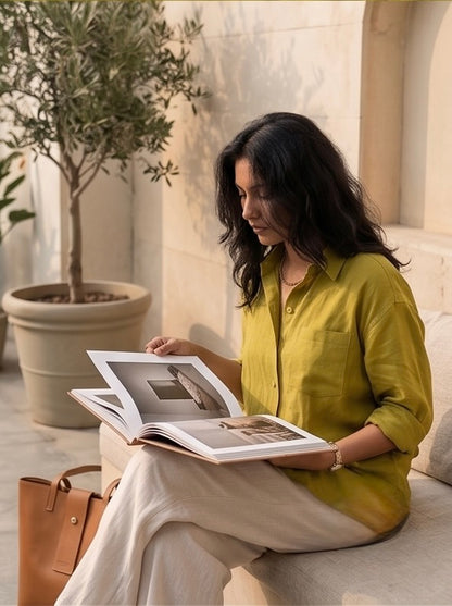 Woman in a green linen shirt reading a book in a cozy indoor setting with a plant and beige walls.